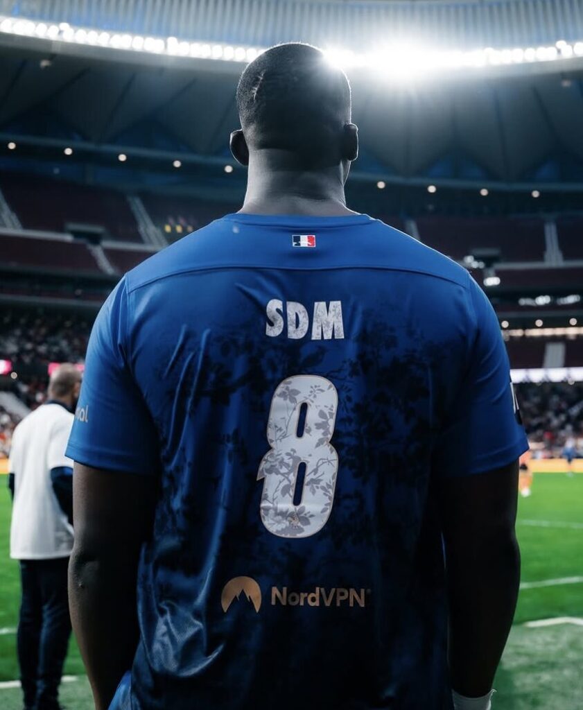 SDM est debout dans un stade, vu de dos. Il porte un maillot avec l’inscription "SDM 8" celui du Stream for Humanity.